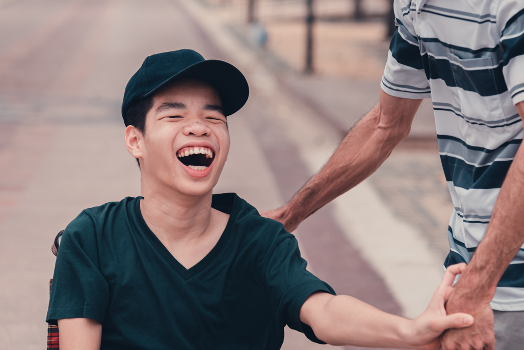 Asian disabled teenager boy on wheelchair and carer in public places, Lifestyle of happy disability child travel holidays at the road in front of beach.